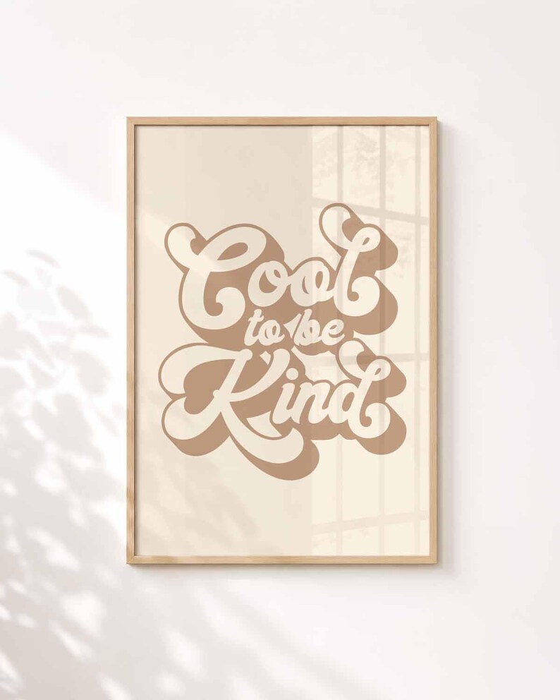 K&ouml;nnte beinhalten: Ein rechteckiger Wandkunstdruck in einem hellen Holzrahmen mit dem Text Cool to be Kind in einer fetten, braunen, retro-inspirierten Schriftart mit Schlagschatteneffekt auf einem einfarbigen, hellbeigen Hintergrund im Rahmen.