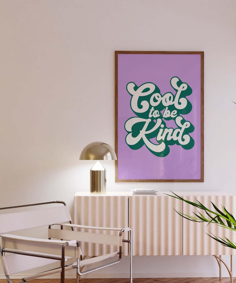 K&ouml;nnte beinhalten: Ein rechteckiger Wandkunstdruck in einem Holzrahmen mit dem Text Cool to be Kind in einer wei&szlig;en, fetten, abgerundeten Schrift mit dunkelgr&uuml;nem Schlagschatten auf einem einfarbigen, hellvioletten Hintergrund, pr&auml;sentiert an einer Wand.