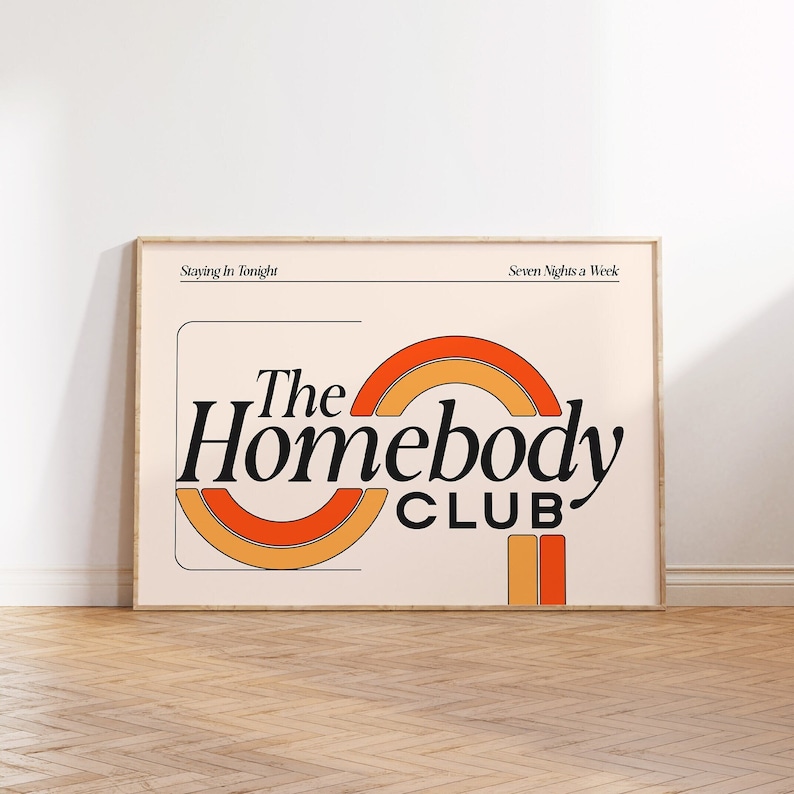 以下が含まれることがあります： 桃色の背景に黒い文字で「The Homebody Club」と書かれた額装されたプリント。テキストはオレンジと黄色の半円形に囲まれています。プリントの上部には「Staying In Tonight」、下部には「Seven Nights a Week」とあります。