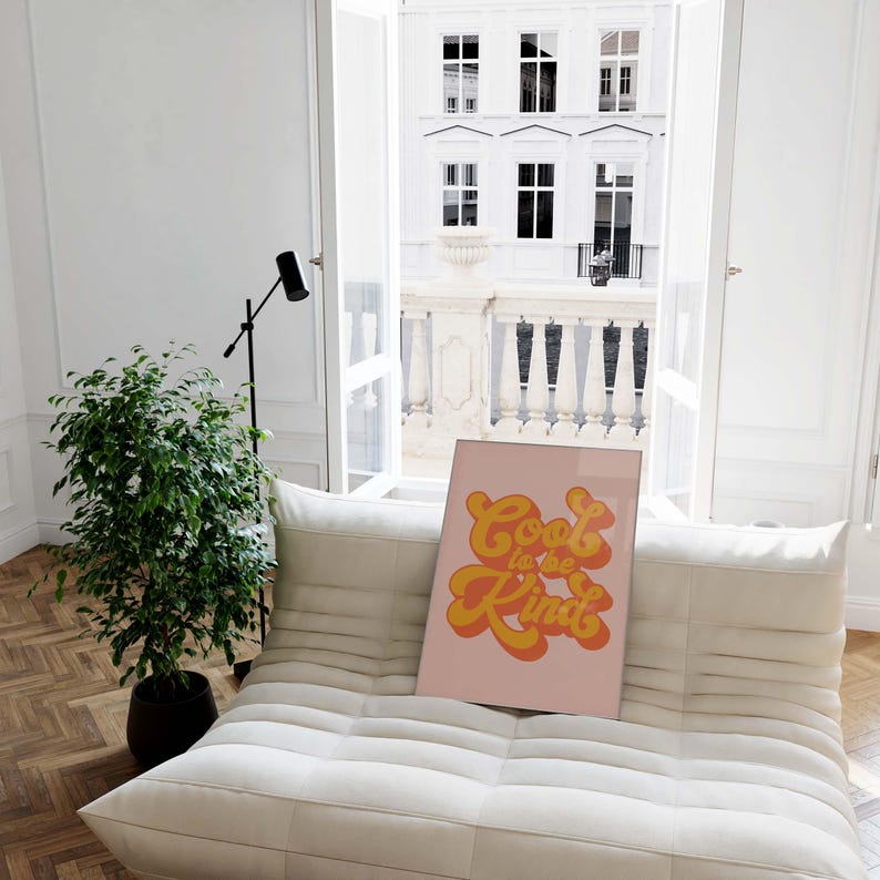 K&ouml;nnte beinhalten: Ein rechteckiger gerahmter Druck mit hellrosa Hintergrund und dem Text Cool to be Kind in einer fetten, orange-gelben Schrift im Retro-Stil. Das Kunstwerk steht auf einem wei&szlig;en, gesteppten Sofa in einem hellen, luftigen Raum.