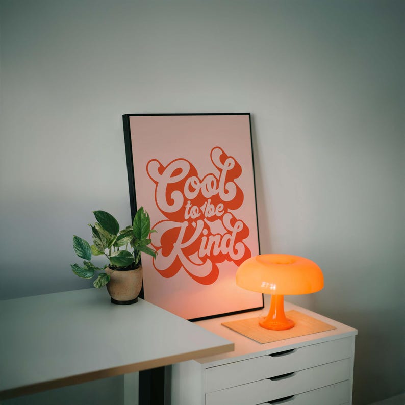 K&ouml;nnte beinhalten: Ein gerahmter rechteckiger Wandkunstdruck mit hellrosa Hintergrund und dem orangefarbenen Text Cool to be Kind in einer fetten, retro-inspirierten Schriftart. Der Druck steht neben einer kleinen gr&uuml;nen Topfpflanze und einer leuchtend orangefarbenen Pilzlampe.