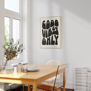 May include: A black and white poster with the text "GOOD VIBES ONLY" in a retro, stylized font. The poster is framed in a thin gold frame and is hanging on a white wall.
