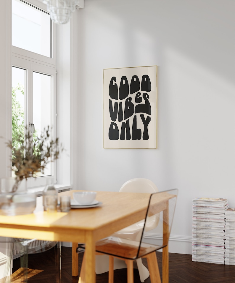 May include: A black and white poster with the text "GOOD VIBES ONLY" in a retro, stylized font. The poster is framed in a thin gold frame and is hanging on a white wall.
