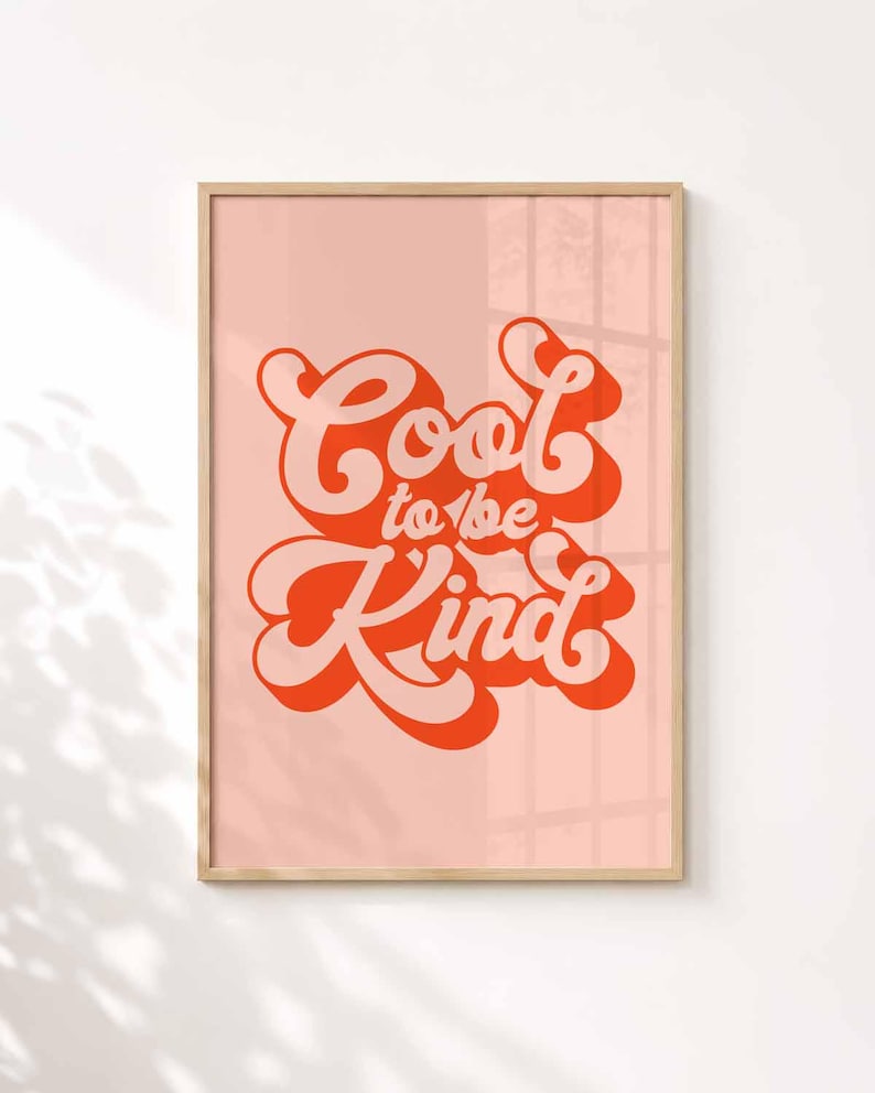 K&ouml;nnte beinhalten: Ein gerahmter Wandkunstdruck mit dem Text Cool to be Kind in einer fetten, stilisierten orangefarbenen Schrift mit einem zartrosa Schlagschatten. Der Schriftzug ist auf einem hellrosa Hintergrund zentriert und von einem hellen Holzrahmen umgeben.