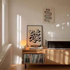 K&ouml;nnte beinhalten: Ein helles Holz-Medienm&ouml;bel mit vertikalen Lamellen, auf dem ein Plattenspieler, eine wei&szlig;e Kugellampe und ein gerahmtes Poster mit dem schwarzen Text Cool to be Kind stehen. Dar&uuml;ber befindet sich ein gerahmter Druck mit einem grauen, geschl&auml;ngelten Pfad.