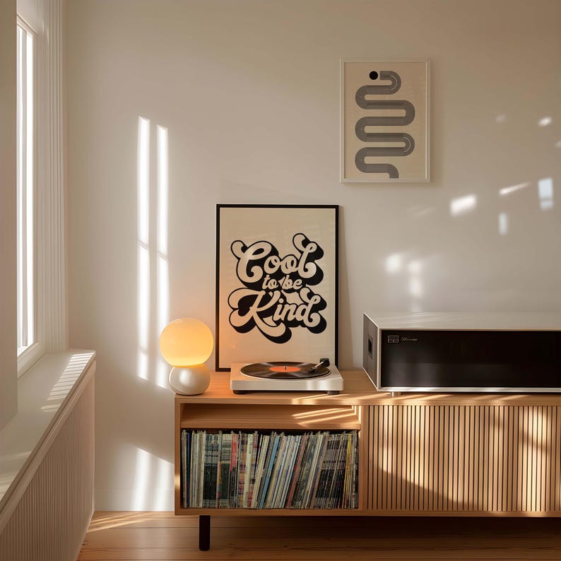 K&ouml;nnte beinhalten: Ein helles Holz-Medienm&ouml;bel mit vertikalen Lamellen, auf dem ein Plattenspieler, eine wei&szlig;e Kugellampe und ein gerahmtes Poster mit dem schwarzen Text Cool to be Kind stehen. Dar&uuml;ber befindet sich ein gerahmter Druck mit einem grauen, geschl&auml;ngelten Pfad.