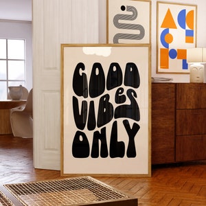 May include: A framed poster with a white background and black text that reads "GOOD VIBES ONLY" in a retro, wavy font. The poster is hanging on a white wall next to two other framed posters.