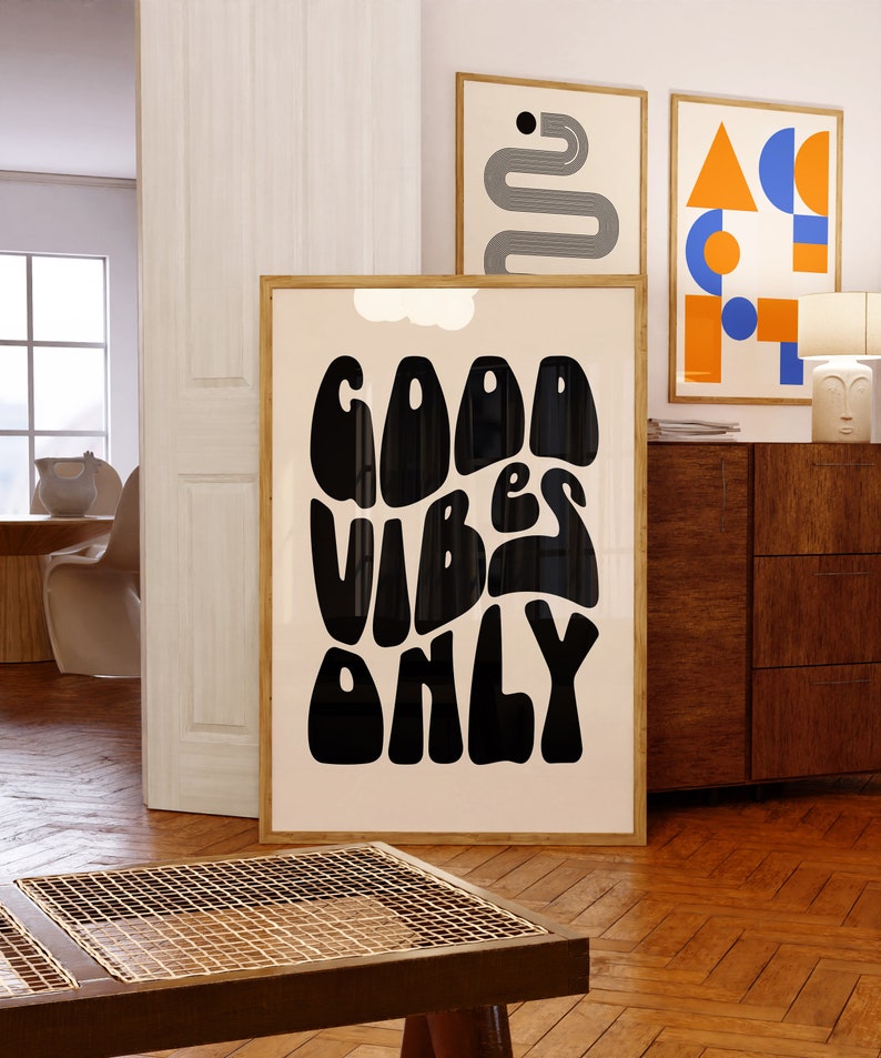May include: A framed poster with a white background and black text that reads "GOOD VIBES ONLY" in a retro, wavy font. The poster is hanging on a white wall next to two other framed posters.