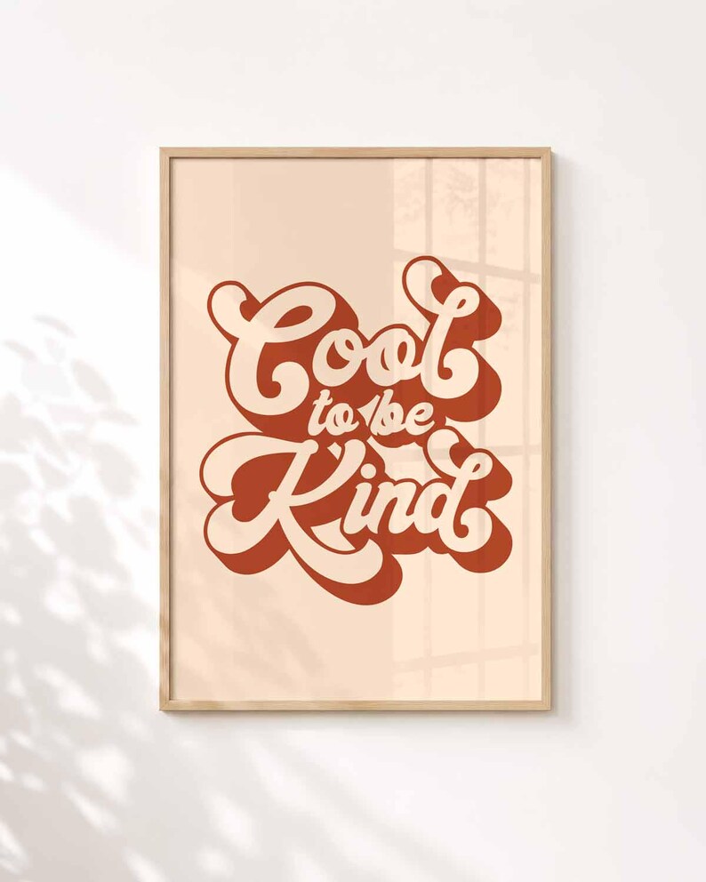 K&ouml;nnte beinhalten: Ein rechteckiger Wandkunstdruck in einem hellen Holzrahmen mit dem Text Cool to be Kind in einer fetten, dunkelorangen Retro-Schriftart. Die Schrift ist mit einem dunkelorangen Schlagschatten auf einem beigen Hintergrund versehen.