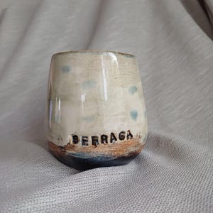 Op de afbeelding: Een handgemaakte keramische beker met een beige glazuur, blauwe hartaccenten en een bruine basis. Het woord "BERRAGA" is in zwarte letters nabij de basis gedrukt. De beker heeft een glanzende afwerking en een organische vorm.