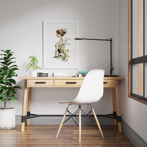 May include: A light wood desk with two drawers, a black desk lamp, and a white chair. A framed watercolor print of a woman with floral accents hangs on the wall. A potted plant sits on the desk, and another is on the floor.