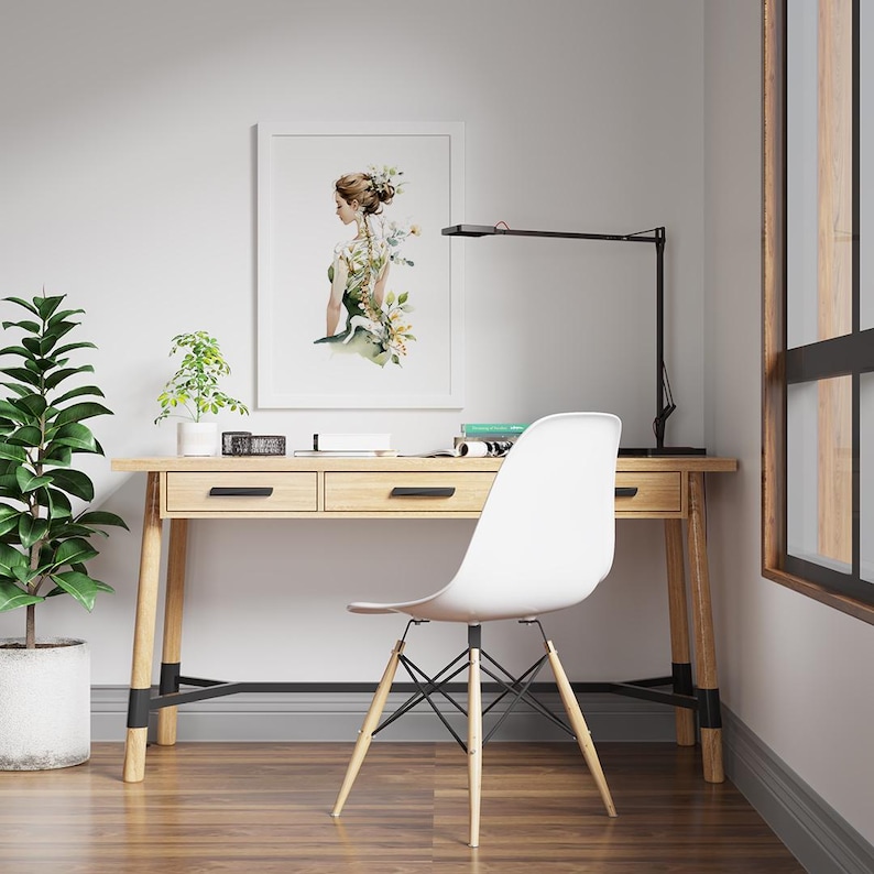 May include: A light wood desk with two drawers, a black desk lamp, and a white chair. A framed watercolor print of a woman with floral accents hangs on the wall. A potted plant sits on the desk, and another is on the floor.
