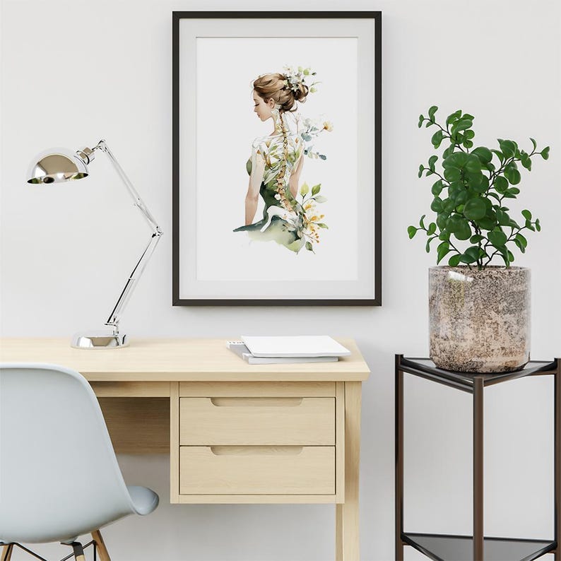 May include: A framed watercolor print of a woman with a floral spine and a green dress. The artwork is displayed on a light wood desk with a silver lamp and a white chair. A potted plant sits on a black side table.