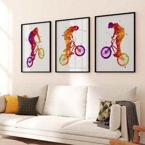 May include: Three watercolor paintings of a person riding a BMX bike. The paintings are in a rainbow of colors, including red, orange, yellow, green, blue, indigo, and violet. The paintings are framed in black frames and are hanging on a white wall.