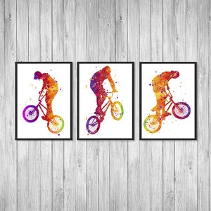 May include: A set of three watercolor prints featuring a silhouette of a person performing a stunt on a BMX bike. The prints are framed in black and have a white background. The colors in the prints are shades of orange, yellow, red, and purple.