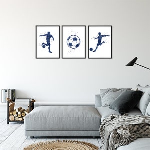 May include: Three framed prints of a soccer player kicking a ball. The prints are in a watercolor style with a blue and white color scheme. The prints are hanging on a white wall above a gray couch.