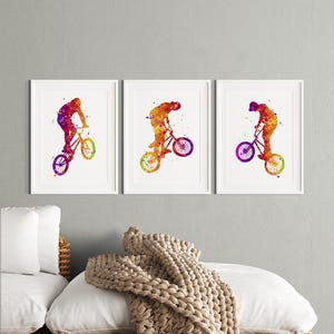 May include: Three watercolor paintings of a person riding a BMX bike. The paintings are in shades of orange, yellow, red, purple, and blue. The paintings are framed in white frames and are hanging on a white wall.