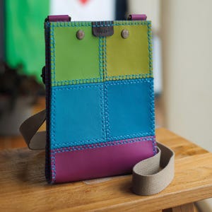 May include: A vibrant leather crossbody bag with a patchwork design. The bag features green, blue, and purple panels, stitched together with blue thread. It has a beige strap and is secured with two metal rivets.