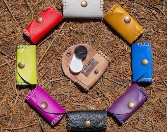 Könnte beinhalten: Eine Ansammlung von acht kleinen, rechteckigen Lederetuis in verschiedenen Farben wie Rot, Gelb, Blau, Lila, Schwarz, Weiß und Limettengrün, jeweils mit einem Messingdruckknopf und sichtbaren Nähten, die ein zentrales, offenes Kontaktlinsenetui umgeben.