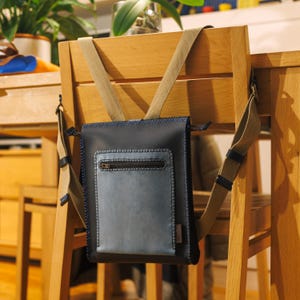 Op de afbeelding: Een blauwe leren tas met een ritsvak en een beige band. De tas hangt aan de rugleuning van een houten stoel.