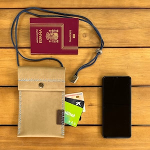 Puede incluir: Una funda de teléfono de cuero marrón con cierre de solapa y un cordón azul. La funda está sobre una superficie de madera con un pasaporte español, un smartphone y tarjetas de crédito.