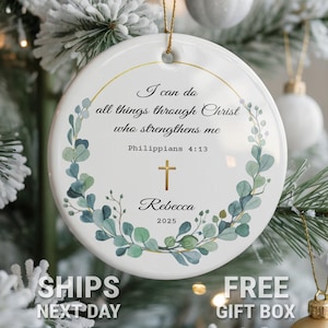 May include: A white ceramic Christmas ornament with a gold-colored rim and a gold string for hanging. The ornament features the text "I can do all things through Christ who strengthens me" and a cross. The name "Rebecca" and the year "2025" are also included.