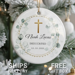 May include: White ceramic Christmas ornament with a gold cross and eucalyptus wreath design. The ornament features the name "Noah Lucas" and the word "Dedicated" with the date "12.10.2025". The ornament is hanging on a Christmas tree.
