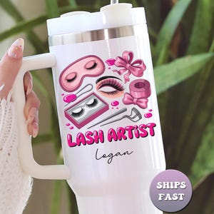 May include: White tumbler with a handle, featuring a pink and black lash artist design. The design includes a pink eye mask, eyelashes, bows, and the words "LASH ARTIST" in pink. The name "Logan" is written below the text.