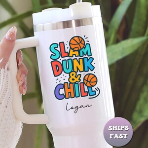 May include: White insulated tumbler with a handle, featuring the text "SLAM DUNK & CHILL" in colorful letters with basketball graphics. The name "Logan" is written below the text. The tumbler has a silver rim and a white lid.