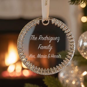 May include: A circular clear glass ornament with a faceted edge hanging from a white ribbon labeled Waterford. The center is etched with the text The Rodriguez Family and Ken, Maria & Hecotr. The ornament hangs from a green pine tree branch.