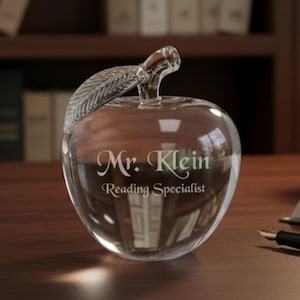 Puede incluir: Un pisapapeles transparente en forma de manzana con un asa en forma de hoja. El texto "Mr. Klein Reading Specialist" est&aacute; grabado en la parte delantera. Un bol&iacute;grafo y un libro abierto est&aacute;n sobre el escritorio de madera.
