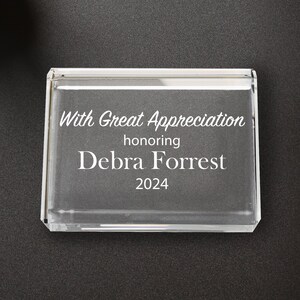 May include: A clear acrylic desk block with the text "With Great Appreciation honoring Debra Forrest 2024". The block is rectangular and sits on a black surface, next to a small plant, a cup of coffee, and a notepad.