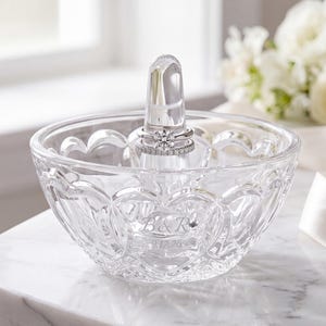 May include: A clear glass ring holder dish with a central cone, featuring an etched design of the letters B & R and the date 9.17.26. The bowl has a scalloped rim and a textured pattern, with a silver-toned ring resting on the central post.