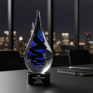 May include: A clear glass award with a teardrop shape, featuring blue and black spiral designs inside. The award sits on a black base with the text "2026 Legacy Award". The background shows a city skyline at night.