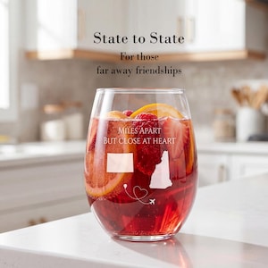 May include: A clear stemless wine glass filled with a red fruit beverage containing orange slices and strawberries. The glass features white etched graphics of two US state outlines, a dashed flight path with a heart, and the text MILES APART BUT CLOSE AT HEART.