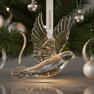 May include: A clear glass bird ornament with wings spread in flight, suspended by a white ribbon printed with the word WATERFORD. The bird features the engraved text Mr. & Mrs. Kline on its side, showcasing detailed feather textures and a polished finish.