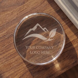 May include: A circular clear glass coaster featuring an etched design of a mountain peak and a single leaf. The text "YOUR COMPANY LOGO HERE" is engraved in the center of the coaster, positioned below the mountain and leaf graphic illustration.