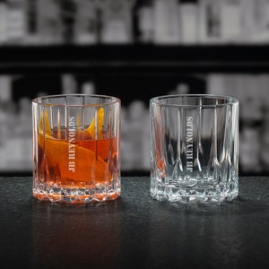 May include: Two clear glass tumblers with a ribbed design. The tumbler on the left contains a reddish-orange liquid and a piece of orange peel. Both tumblers have the text "JB REYNOLDS" etched on the side.