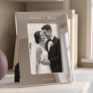 May include: A polished silver-toned rectangular photo frame featuring an engraved inscription at the top reading Danielle & Shawn and a date reading 11.14.26 at the bottom. The frame holds a black and white photograph of a couple in wedding attire.