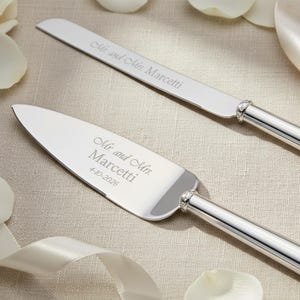 K&ouml;nnte beinhalten: Ein silbernes Tortenmesser- und Tortenheber-Set mit polierten Metallgriffen und einem dekorativen Band mit Kristallbesatz. Das Messer ist mit Mr. and Mrs. Marcetti graviert, der Heber mit Mr. and Mrs. Marcetti und 4-10-2026.