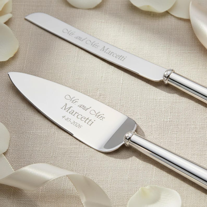K&ouml;nnte beinhalten: Ein silbernes Tortenmesser- und Tortenheber-Set mit polierten Metallgriffen und einem dekorativen Band mit Kristallbesatz. Das Messer ist mit Mr. and Mrs. Marcetti graviert, der Heber mit Mr. and Mrs. Marcetti und 4-10-2026.