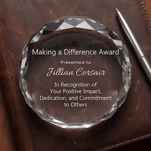 May include: A circular, faceted clear glass award featuring the engraved text: Making a Difference Award, Presented to Jillian Corsair, In Recognition of Your Positive Impact, Dedication, and Commitment to Others. A silver pen is positioned nearby.