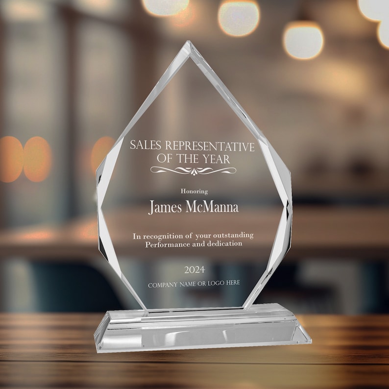 Puede incluir: Un premio de cristal transparente en forma de diamante con el texto "SALES REPRESENTATIVE OF THE YEAR" y "Honoring James McManna". El premio tambi&eacute;n incluye el texto "In recognition of your outstanding Performance and dedication" y "2024 COMPANY NAME OR LOGO HERE".