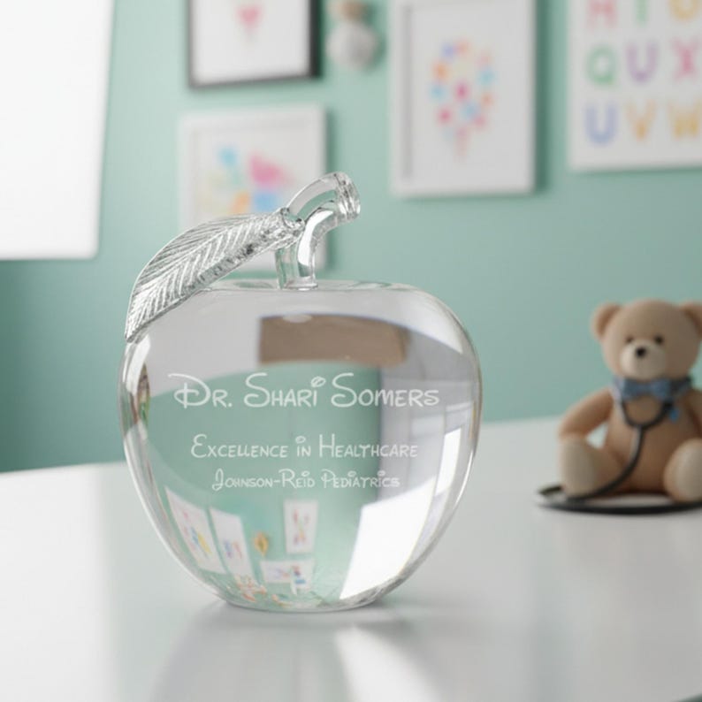 Puede incluir: Una pieza decorativa en forma de manzana de cristal transparente con un tallo de hoja. La manzana tiene grabado el texto: "Dr. Shari Somers, Excellence in Healthcare, Johnson-Reid Pediatrics." Un oso de peluche con un estetoscopio est&aacute; cerca.