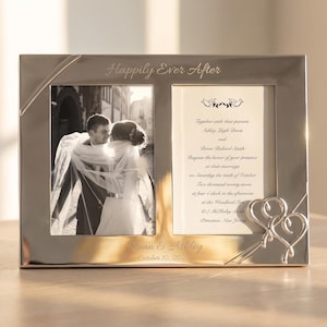 May include: A silver-toned rectangular double photo frame featuring the engraved text "Happily Ever After" at the top and "Brian & Ashley October 10, 2027" at the bottom. The frame includes two metal heart accents and holds a wedding photo and invitation.