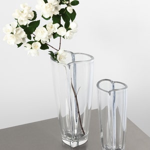 Op de afbeelding: Twee heldere glazen vazen met een hartvormige bovenkant en een geometrische basis. E&eacute;n vaas bevat een tak met witte bloemen met groene bladeren. De vazen staan op een grijs oppervlak tegen een witte achtergrond.