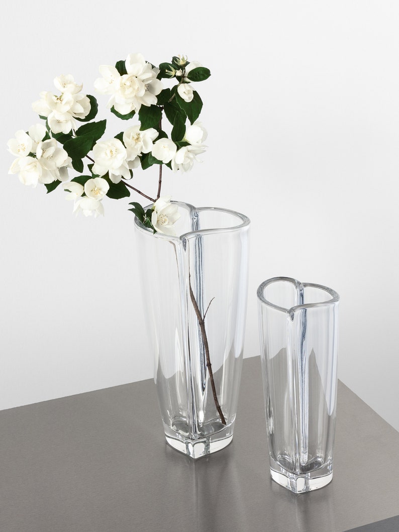 Op de afbeelding: Twee heldere glazen vazen met een hartvormige bovenkant en een geometrische basis. E&eacute;n vaas bevat een tak met witte bloemen met groene bladeren. De vazen staan op een grijs oppervlak tegen een witte achtergrond.