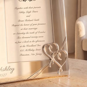 May include: A silver-toned metal picture frame featuring two intertwined heart accents on the lower right corner. The frame displays a printed wedding invitation for Ashley Leigh Davis and Brian Richard Smith, dated October 10, 2027, at the Woodland Inn.