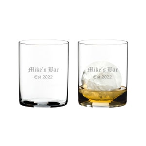May include: Two clear glass tumblers with the text "Mike's Bar Est 2022" etched on the side. One tumbler is empty and the other has a large ice sphere and amber liquid.