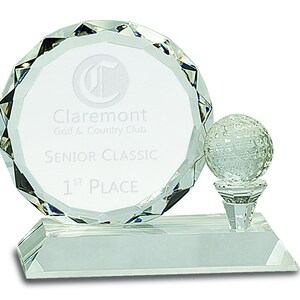 Puede incluir: Un trofeo de golf de cristal transparente con una placa redonda que dice "Claremont Golf & Country Club Senior Classic 1st Place". Una bola de golf se sienta en la parte superior de un tee.
